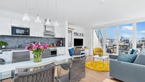Un appartement moderne comprenant une cuisine, un coin repas et un salon. Il dispose d’une grande fenêtre offrant une vue sur la ville, de meubles en osier et d’un tapis jaune.