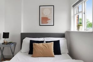 A bedroom with a bed, pillows, a bedside table with a lamp, and a window with a view of greenery.