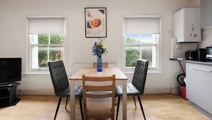 A modern kitchen with a dining area, featuring a wooden table, two chairs, a vase with flowers, and a view of greenery through the windows. - Crouch Hill N4, Crouch Hill, Finsbury Park (London)