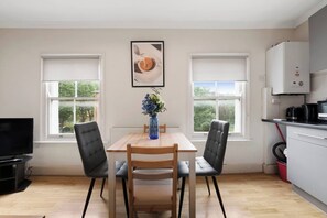 A modern kitchen with a dining area, featuring a wooden table, two chairs, a vase with flowers, and a view of greenery through the windows.