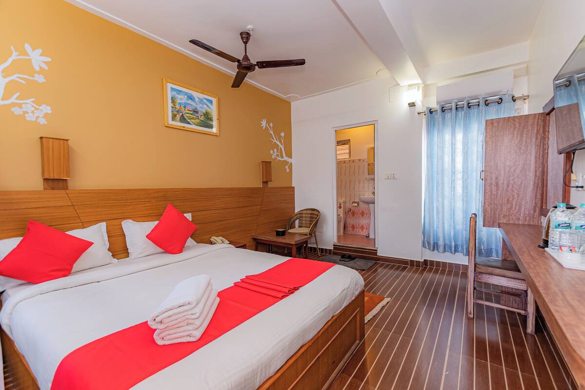A hotel room with a wooden bed, red pillows, a ceiling fan, a painting on the wall, and a wooden floor.
