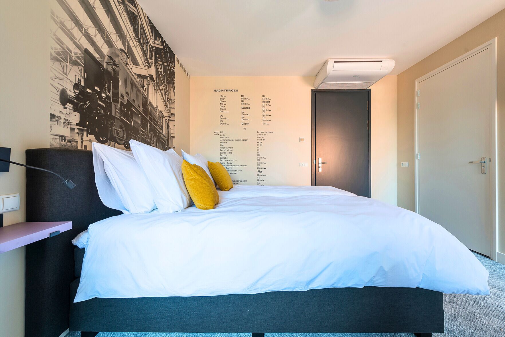 Hotellihuone, jossa on suuri sänky, tumma pääty, seinälle asennettu ilmastointilaite ja ovi oikealla puolella.