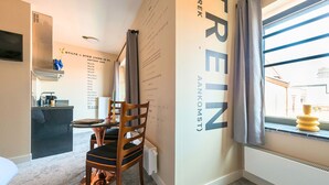Una habitación de hotel moderna con zona de comedor, cocina y un amplio ventanal con cortinas.