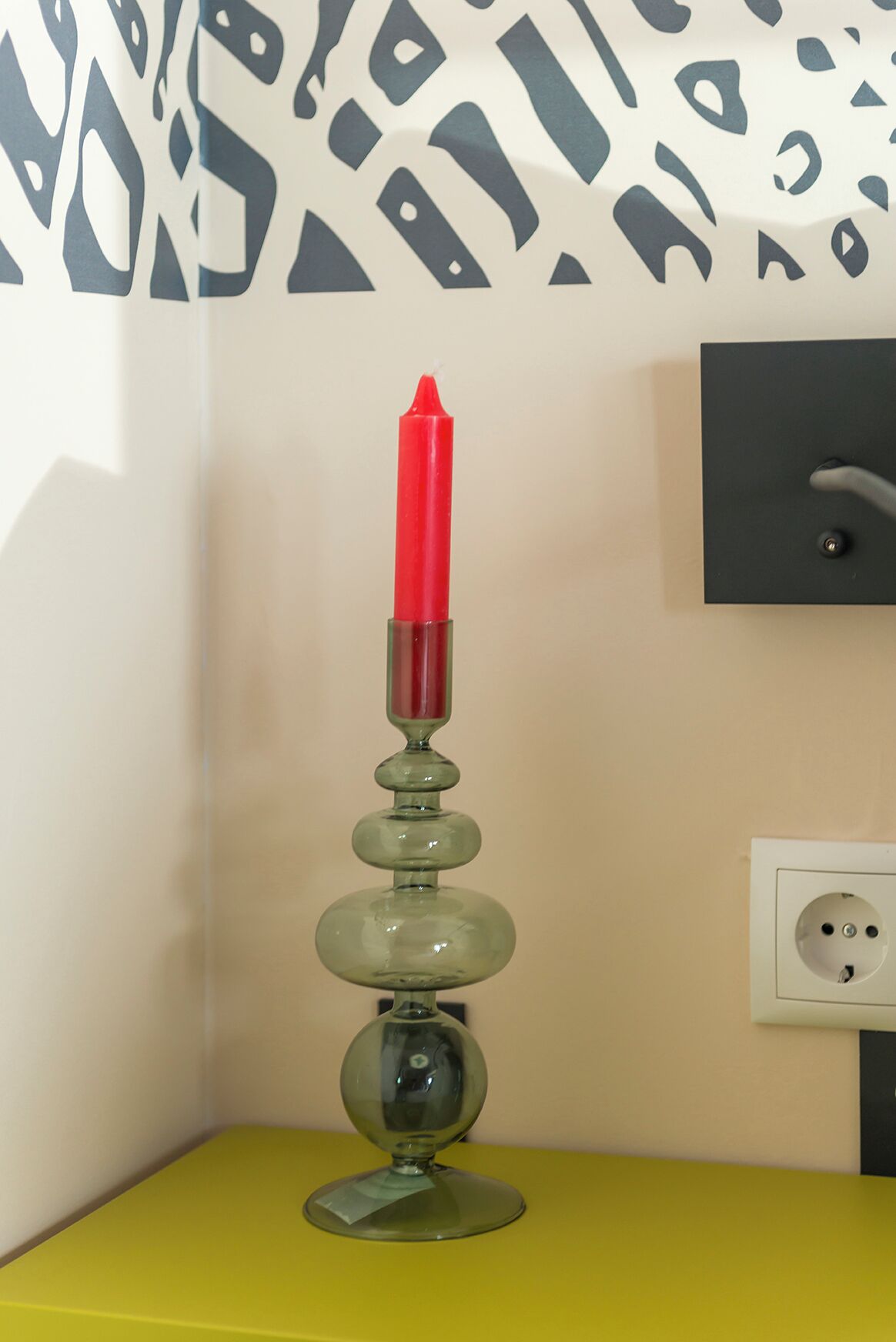 A red candle on a green glass holder on a yellow table against a wall with a black and white abstract pattern.