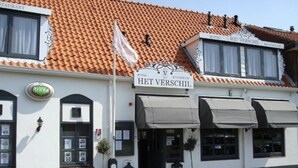 A white building with a tiled roof, a sign reading "HET VERSCHIL HOTEL RESTAURANT," and a flag on top.