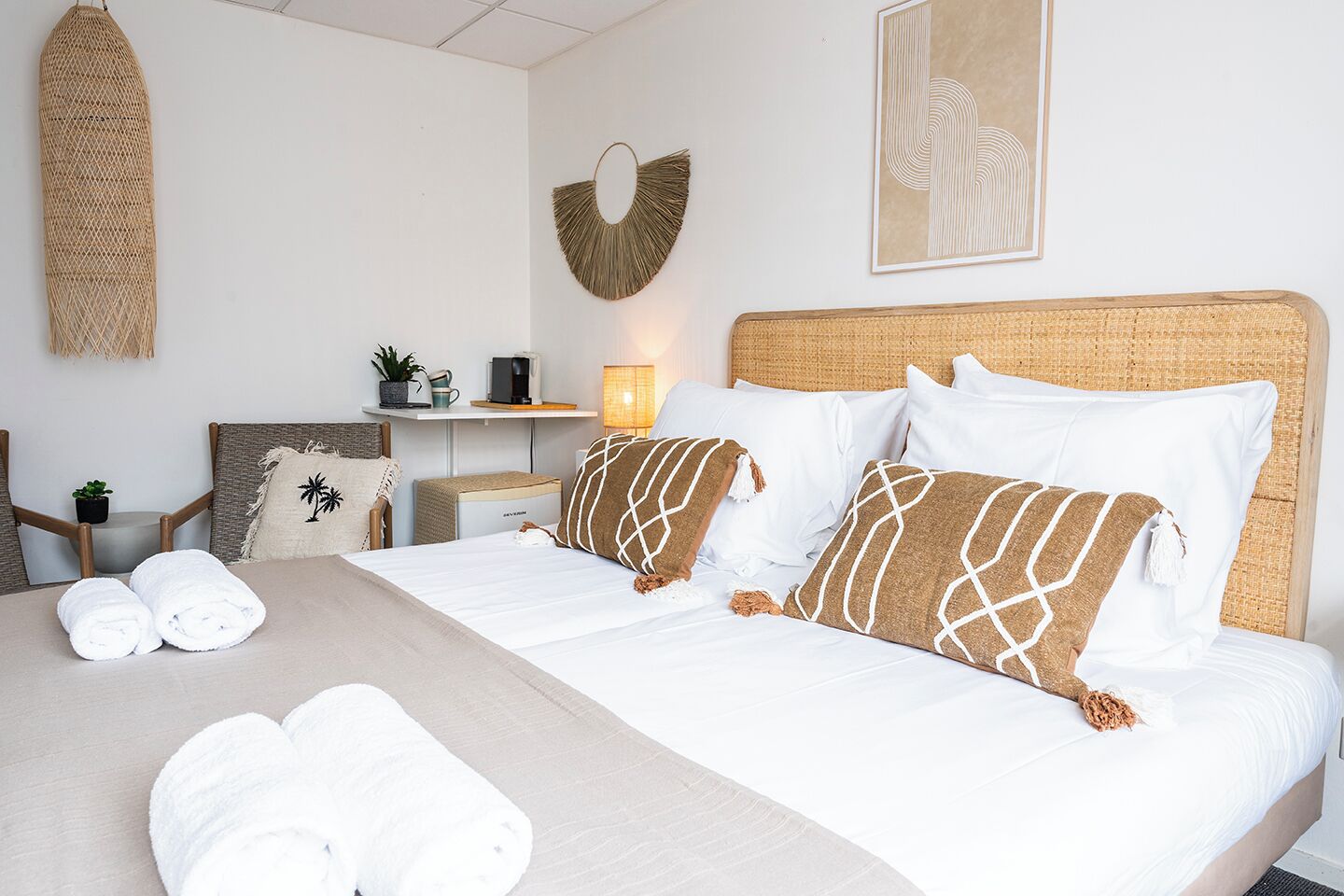 A hotel room with a bed, two pillows, a bedside table with a lamp, a small plant, and a wall-mounted decorative item.