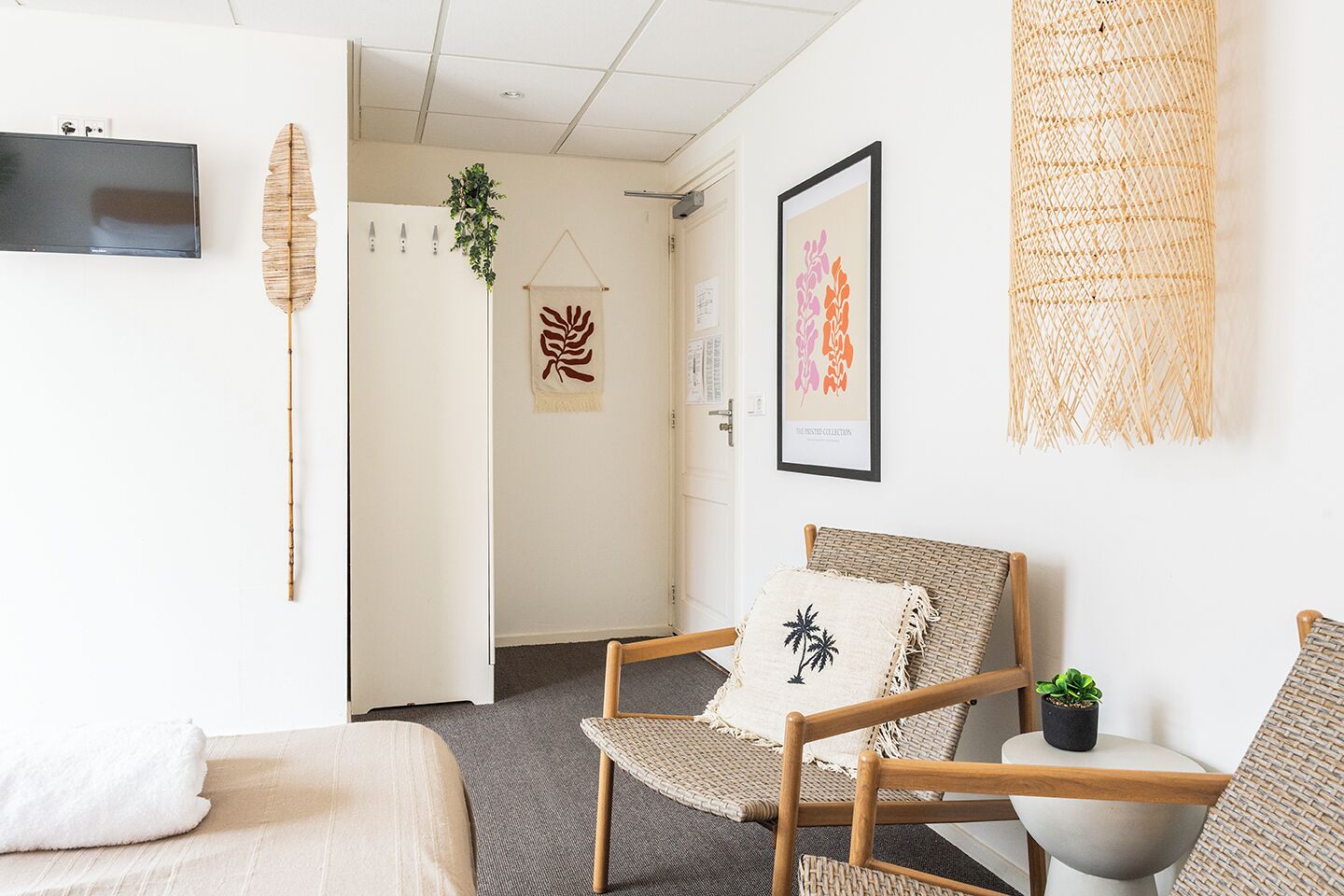 Una habitación de hotel moderna con una cama, un sillón de mimbre, una mesita con una planta y un televisor montado en la pared.