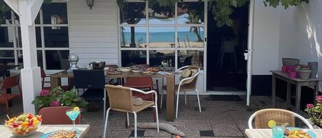 An outdoor patio area with tables and chairs, a pergola with draped fabric, and a view of the beach through large windows.