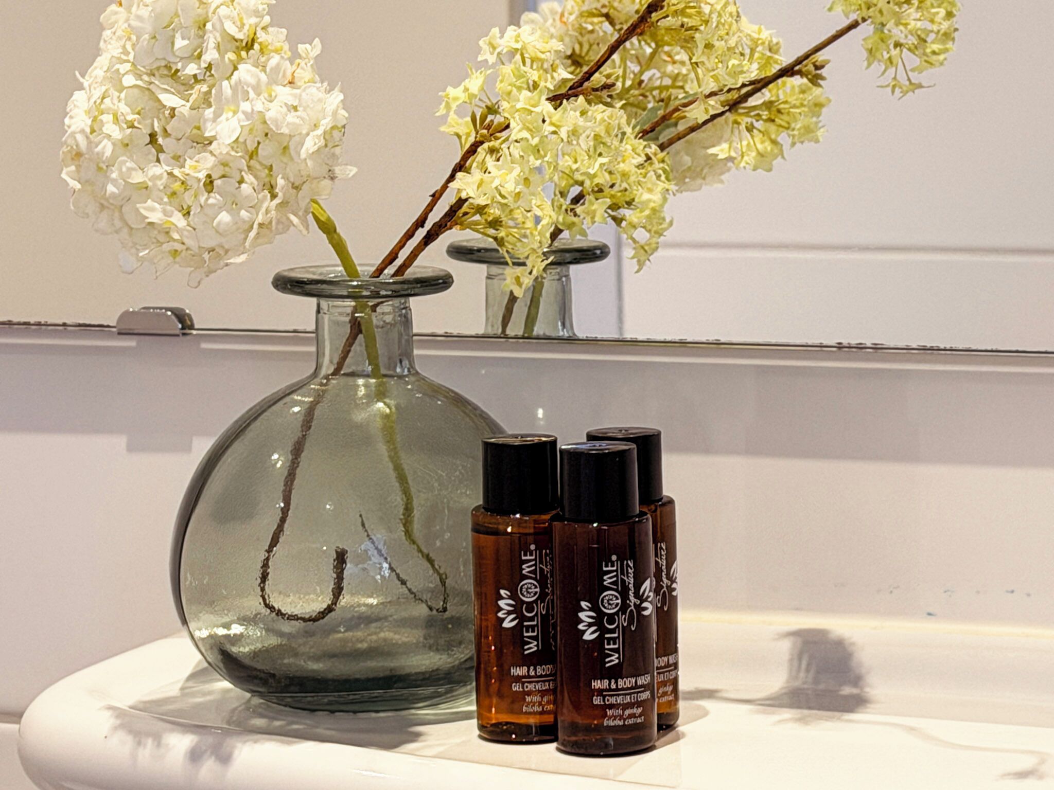 Un florero de vidrio transparente con flores blancas, un espejo y tres botellas de productos para el cabello y el cuerpo sobre una superficie blanca.