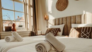 A hotel room with two beds, a window with a view of buildings, a bedside table with a vase, and a wall-mounted lamp.