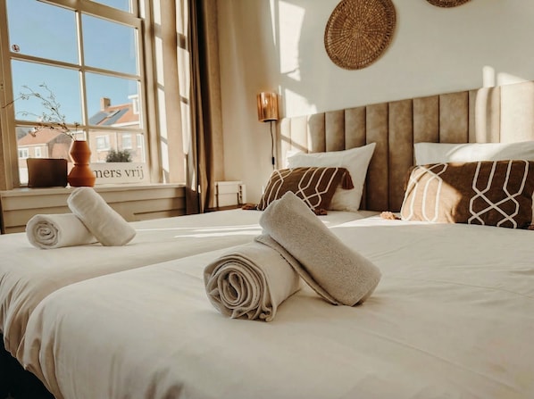 Habitación de hotel con dos camas, una ventana que ofrece vistas a edificios, una mesita de noche con un florero y una lámpara de pared.