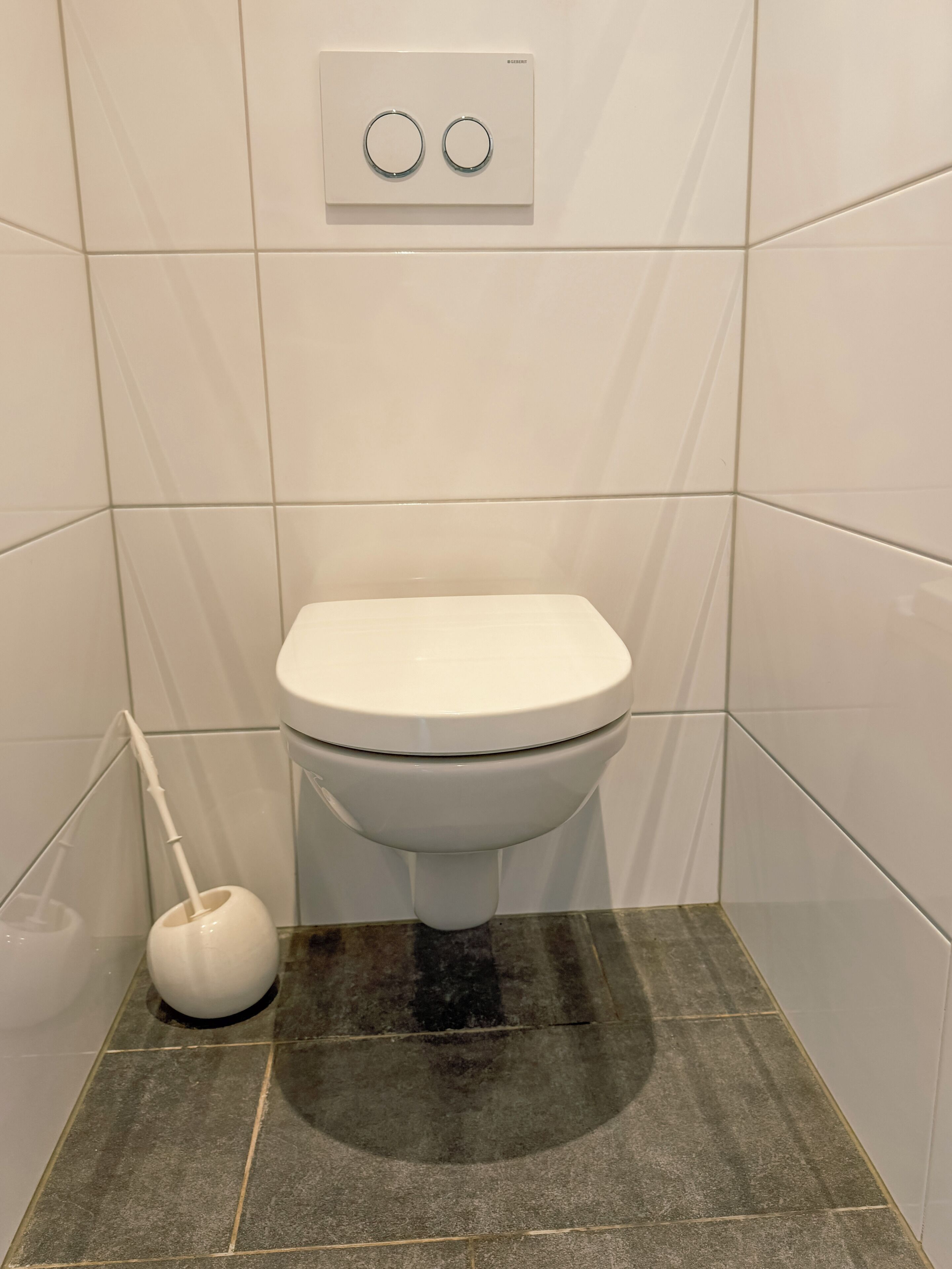 Een wit toilet met een spoelhendel, een zuiger en een bedieningsknop aan de muur.