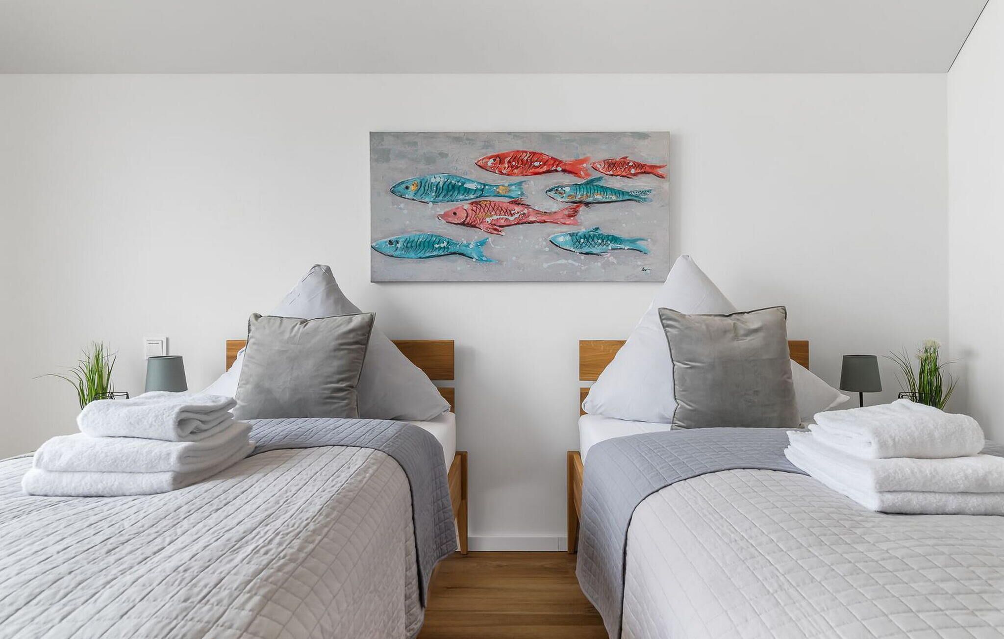 Une chambre d’hôtel avec deux lits, chacun étant équipé d’un oreiller et d’une serviette pliée. Un tableau représentant des poissons est accroché au mur, au-dessus des lits.