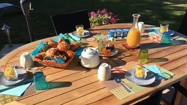 Une table ronde en bois, mise pour le petit-déjeuner, avec des croissants, une théière et diverses boissons.