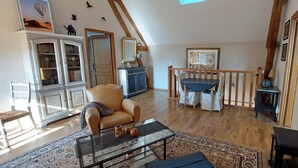 Un vaste salon doté d’un haut plafond, de poutres apparentes, d’un coin repas avec une table et des chaises, et d’une cheminée.