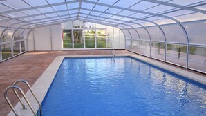 An indoor swimming pool with a glass ceiling, surrounded by a white wall and a wooden deck.
