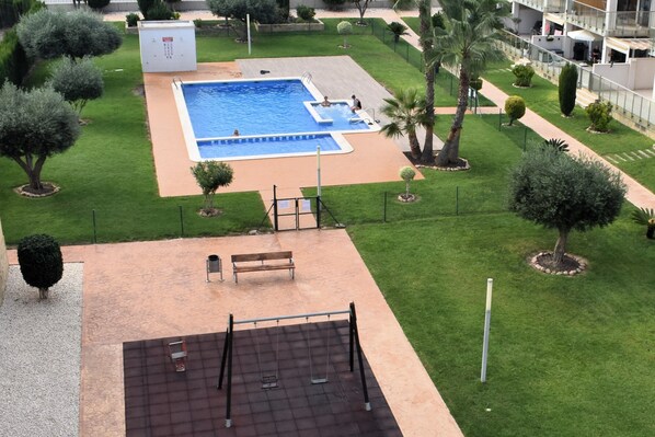 Una piscina rodeada de césped cuidado, un banco y una zona de juegos.