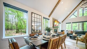 Ein Essbereich mit großem Fenster zum Wald, einem Kamin und einer gemütlichen Sitzecke mit Sofa und Sesseln.