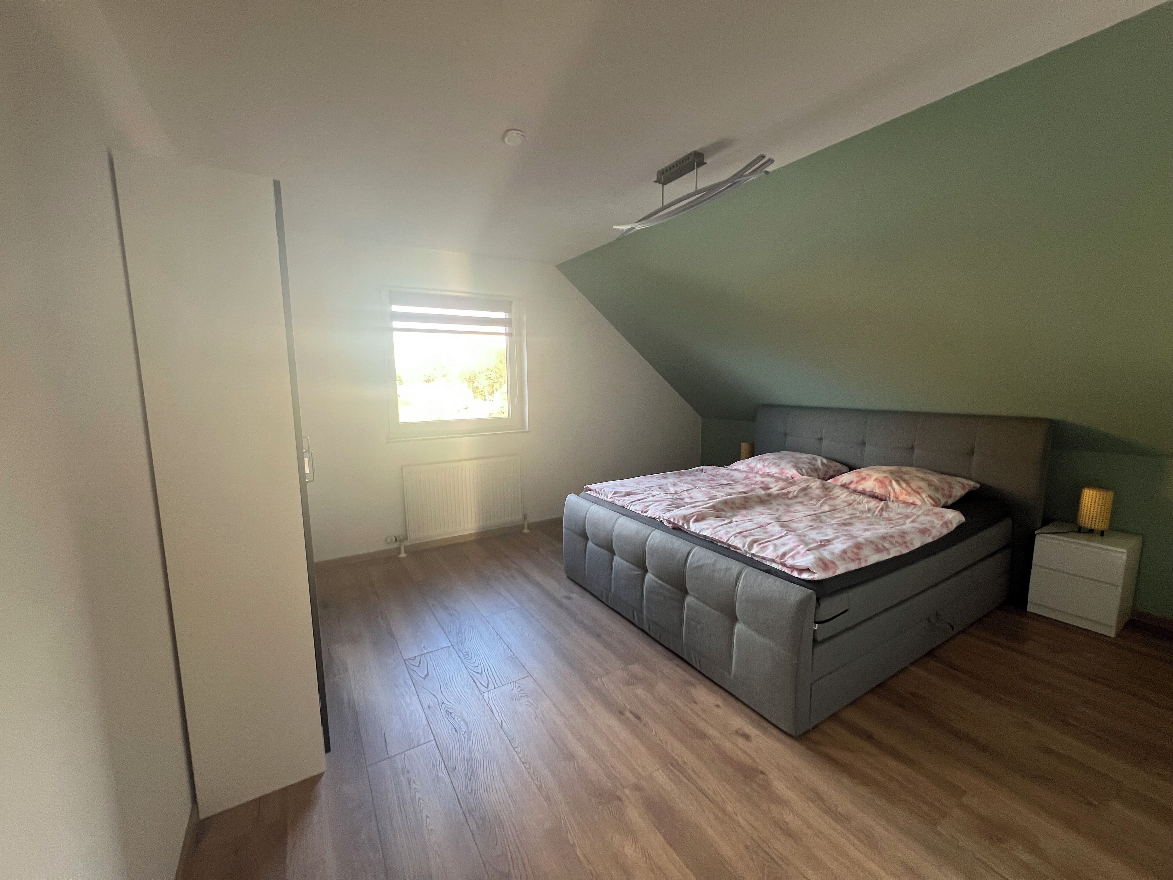A bedroom with a sloped ceiling, a bed with a grey headboard, a bedside table with a lamp, and a window with blinds.