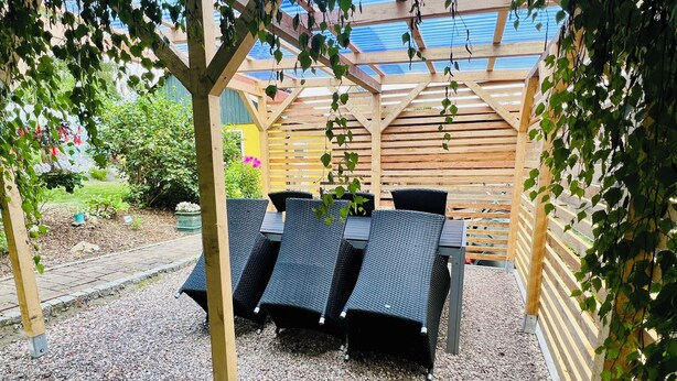 A covered outdoor seating area with a table and chairs, surrounded by gravel and greenery. - Ferienwohnung im Grünen (Hainichen)