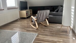 A modern living room with a sloped ceiling, a wooden floor, a grey sofa, a TV on a wooden stand, and a rocking chair. - Ferienwohnung im Grünen (Hainichen)