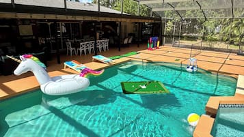 Une piscine extérieure avec un flotteur en forme de licorne, un petit espace de jeux et un coin salon couvert avec des tables et des chaises.