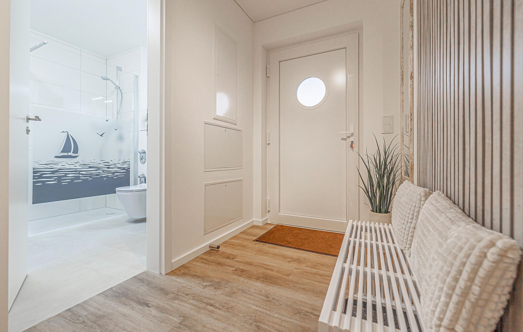 A hallway with a wooden floor, a white bench, a round door, and a bathroom with a sailboat mural.