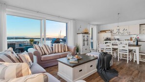 En rummelig stue med et stort vindue der kigger ud over vandet, et hyggeligt siddeområde og et moderne køkken med hvide skabe.