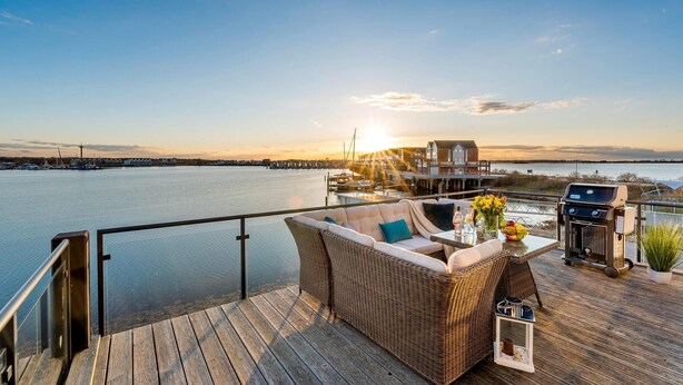 En terrasse med fletmøbler, grill og udsigt over en havn ved solnedgang.