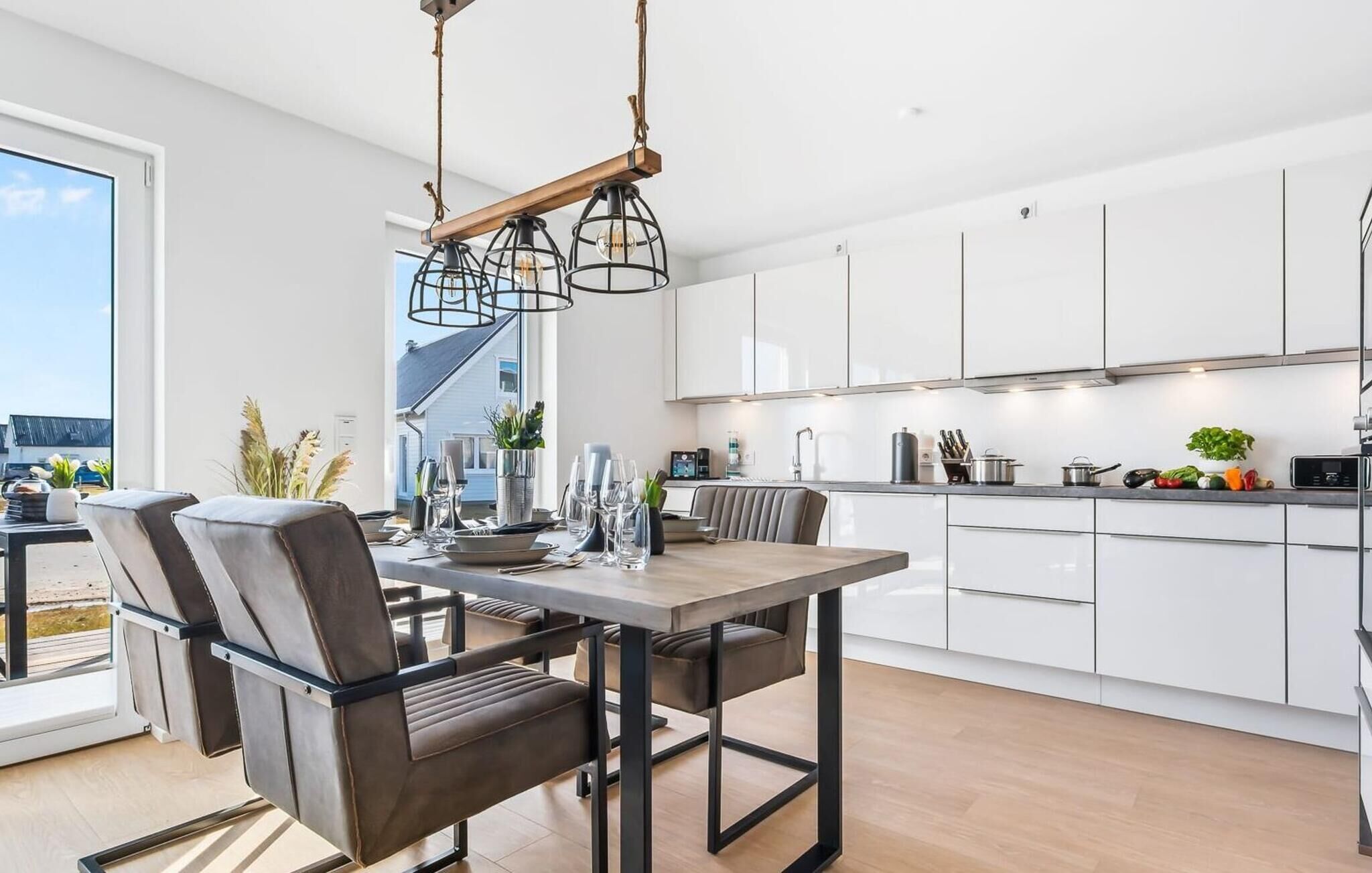A modern kitchen with white cabinets, a dining area with a wooden table and chairs, and a view of the outdoors through a large window.