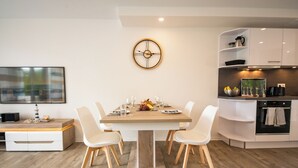 A modern kitchen with a dining table set for four, white cabinetry, and a wall-mounted clock. - Baltic Village - Comforthaus (Damp)