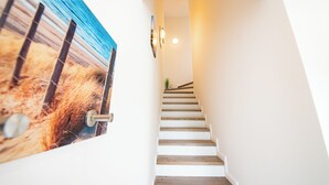 A staircase with white steps leading to a landing, a wall-mounted picture of a beach scene, and a light fixture on the wall. - Baltic Village - Comforthaus (Damp)