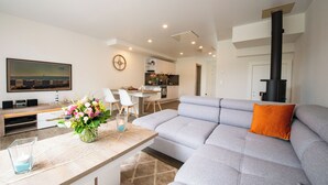 A modern living room with a grey sofa, a wooden coffee table with a vase of flowers, a flat-screen TV, and a kitchen in the background. - Baltic Village - Comforthaus (Damp)