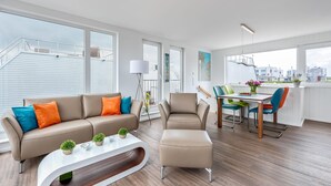 A modern living room with a sofa, ottoman, and coffee table, a dining area with a table and chairs, and large windows offering a city view. - Stunning ship in Olpenitz with sauna (Olpenitz)
