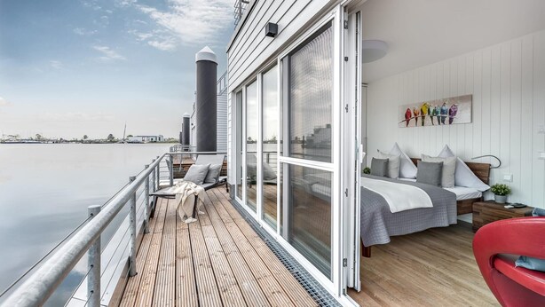 A modern room with a large window, a bed with pillows, a red chair, and a view of the water. - Stunning ship in Olpenitz with sauna (Olpenitz)