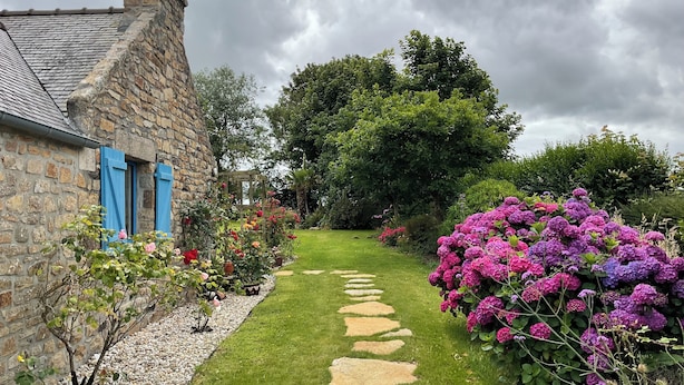 Un bâtiment en pierre avec des volets bleus, un chemin en pierre et un jardin parsemé de fleurs roses.