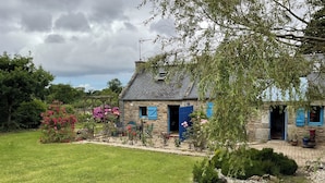 Une maison en pierre avec un toit en pente, un jardin orné de plantes en fleurs et un ciel dégagé.