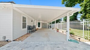 A covered patio with a table and chairs, a white fence, and a pathway leading to a door.
