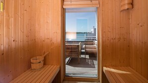 A wooden sauna with a bench and a bucket, a glass door, and a view of a body of water and buildings.