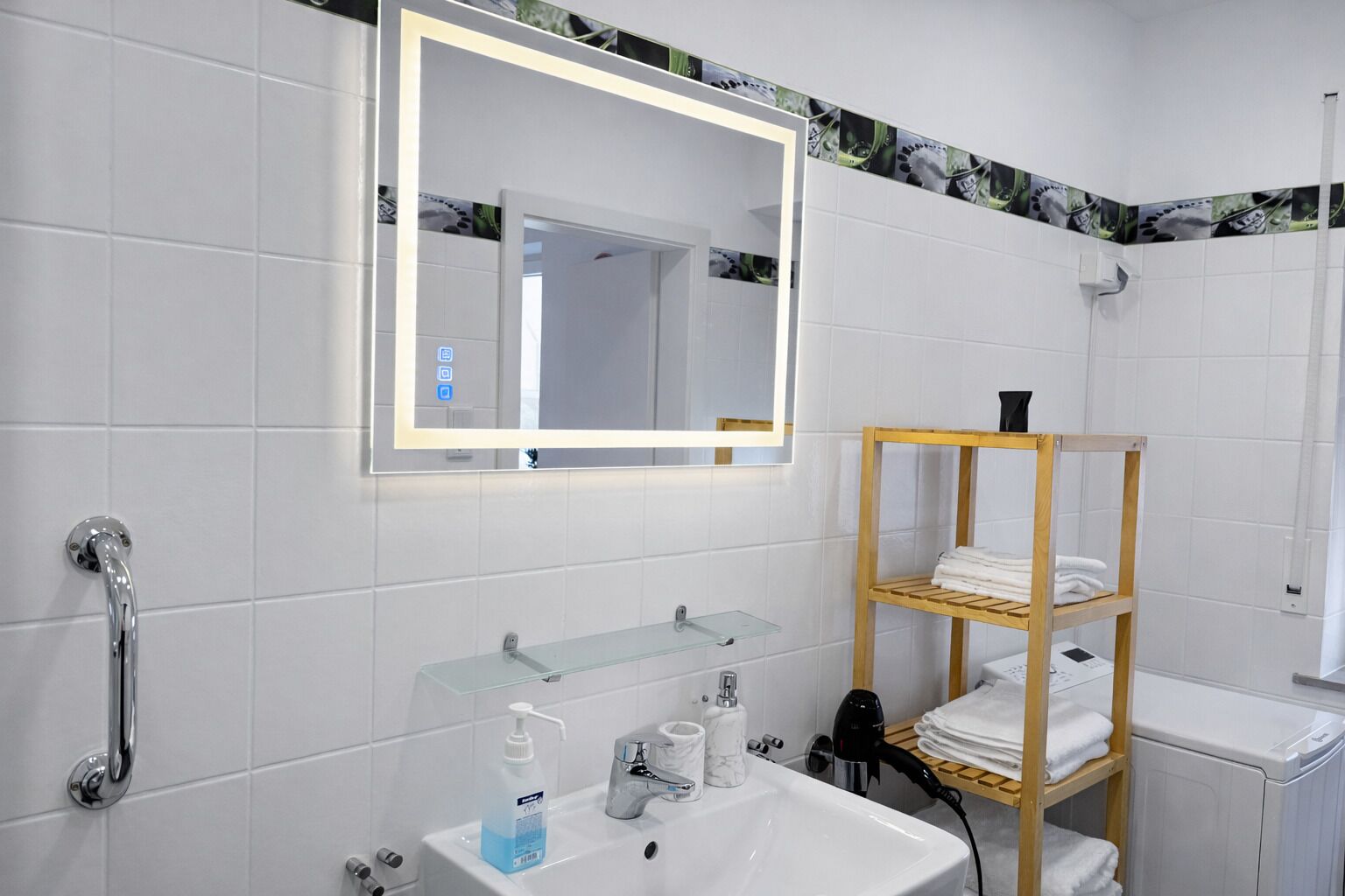 Un baño moderno con estante de vidrio, toallero de madera, lavabo blanco y espejo con marco iluminado.