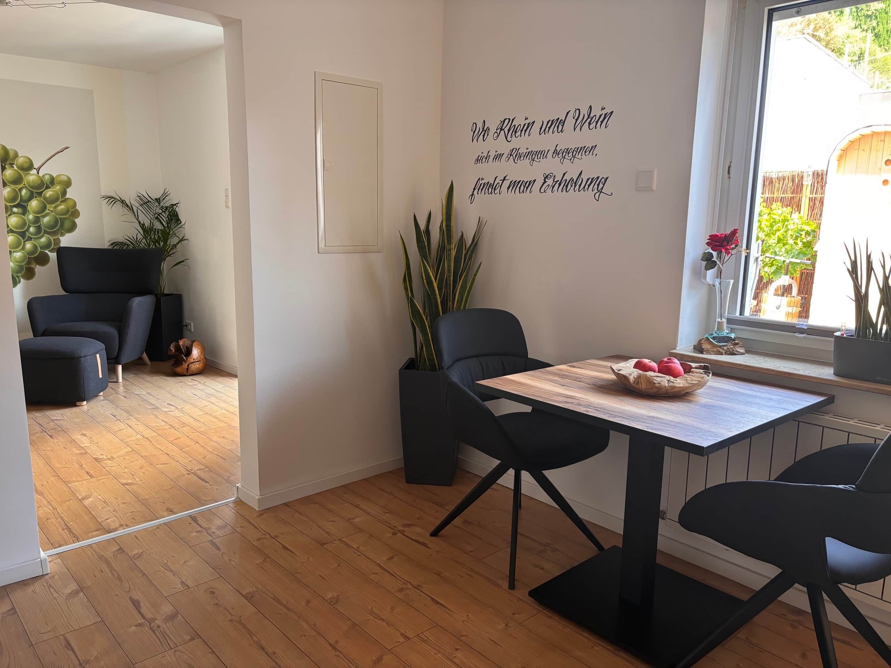 Ein modernes Interieur mit Essbereich, der einen Holztisch und schwarze Stühle, eine Wand mit einer dekorativen Beschriftung sowie einen Blick ins Freie durch ein Fenster zeigt.