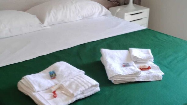 Ein ordentlich gemachtes Bett mit einer grünen Tagesdecke, zwei gefaltete weiße Handtücher und ein weißer Nachttisch mit einer Lampe.