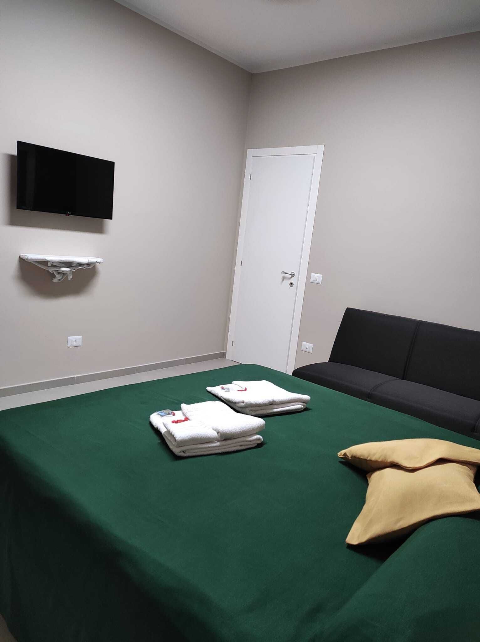 A room with a bed, a TV mounted on the wall, a white door, and a small shelf.