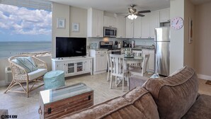 A modern kitchen with white cabinets, a stainless steel refrigerator, and a flat-screen TV. There is a dining area with white chairs and a table, and a living room with a brown sofa and a glass coffee table. - LIFE IS BETTER AT THE BEACH! ESTERO BEACH & TENNIS CLUB 603B! (Fort Myers Beach)