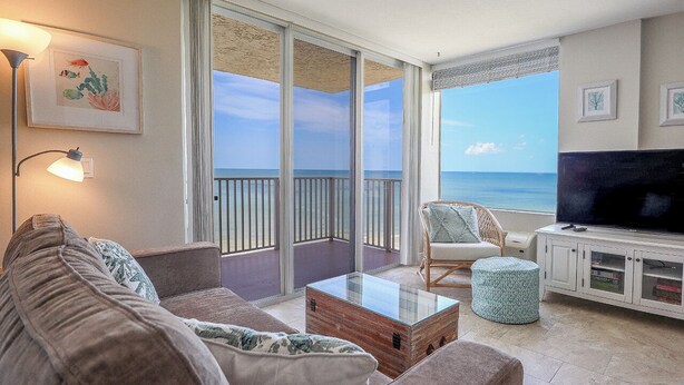 A modern living room with a large TV, comfortable seating, and a balcony with a view of the ocean. - LIFE IS BETTER AT THE BEACH! ESTERO BEACH & TENNIS CLUB 603B! (Fort Myers Beach)