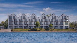 Un immeuble résidentiel moderne à plusieurs étages, doté de balcons, situé près d’un plan d’eau.