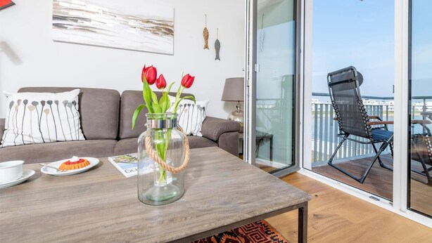 Un salon moderne avec un canapé, une table basse ornée d’un vase de tulipes rouges, et une vue sur un balcon avec un fauteuil.