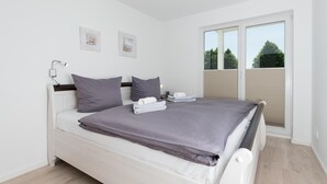 A modern bedroom with a white bed, grey bedding, and a large window with blinds.