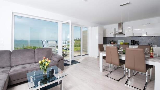 A modern living room with a glass coffee table, grey sofa, and a view of the sea through large sliding doors.