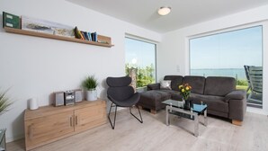 A modern living room with a sofa, a chair, a coffee table, and a bookshelf. There is a large window with a view of the outdoors.